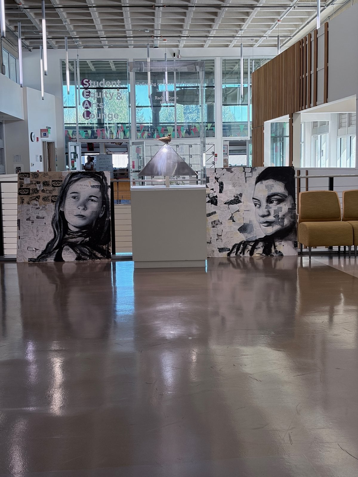 The Rachel Corrie memorial sculpture in its case, displayed between portraits of Rachel Corrie and Ayşenur Ezgi Eygi by Diana Fakhoury and Brady Black in the Student Equity & Arts Lounge at The Evergreen State College
